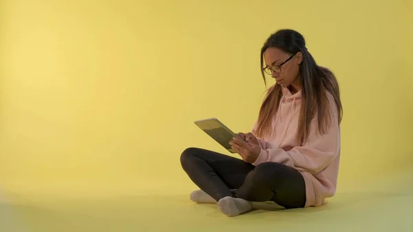 Atractiva chica de raza mixta leyendo algo en la tableta y luego mirando por encima de sus gafas a la cámara — Foto de Stock