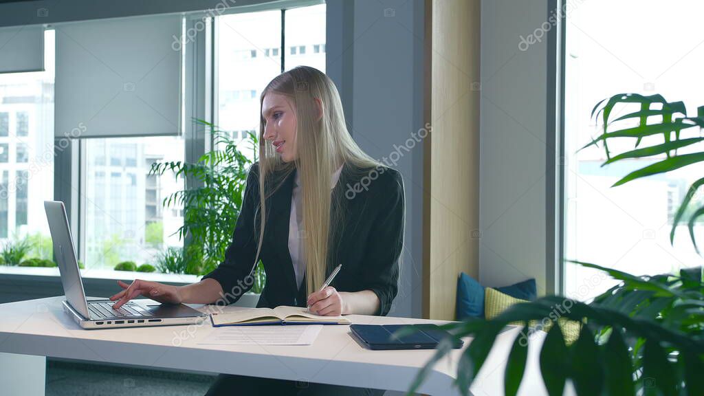 Elegant woman working in stylish office. Modern blond woman in trendy suit sitting at table in light contemporary office and writing on papers.