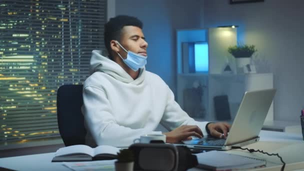 Hombre multiétnico cansado con máscara protectora trabajando en casa hasta tarde en la noche y haciendo actividades de calentamiento — Vídeos de Stock