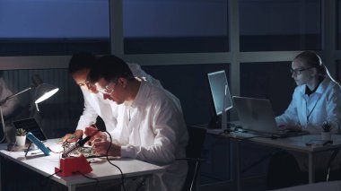 Çok ırklı erkek ve kadın elektronik laboratuarındaki çoklu ölçüm cihazıyla ana kartı kontrol ediyorlar.