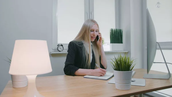 Mujer de negocios hablando por teléfono inteligente en la oficina. Elegante joven rubia en traje de oficina sentada en el lugar de trabajo y negociando a través del teléfono móvil en mano anotando la información necesaria en el cuaderno — Foto de Stock