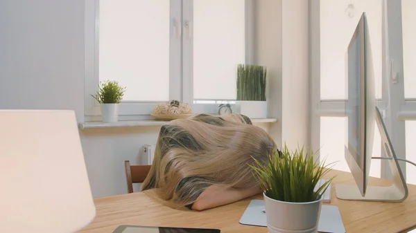 Mujer cansada despertando en el lugar de trabajo. Trabajadora de oficina rubia cansada en traje elegante relajada tumbada en los brazos en el escritorio luego levantarse bostezando y comenzar a trabajar en la computadora en la sala de luz con plantas . — Foto de Stock