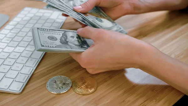 Mani femminili contando contanti con bitcoin. Vista ritagliata delle mani delle donne che contano un grande fascio di banconote da un dollaro su una scrivania di legno con bitcoin e tastiera . — Foto Stock