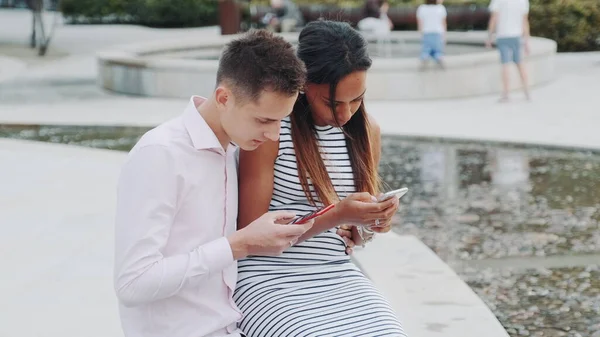 Багатоетнічна пара сидить разом на відкритому повітрі і дивиться в смартфонах — стокове фото