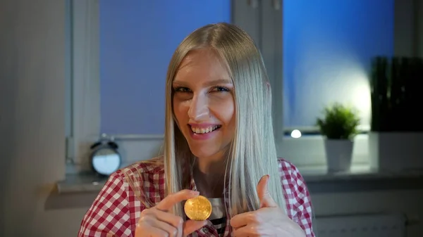 Happy woman holding bitcoin showing thumbs up. Cheerful young blond woman in casual clothes sitting at dark window at night holding bitcoin and doing thumbs up gesture smiling and looking at camera. — Stock Photo, Image