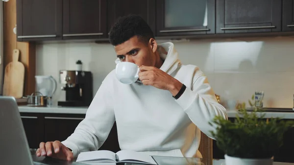 Mixed-race man speaking by smartphone and working on computer at home kitchen