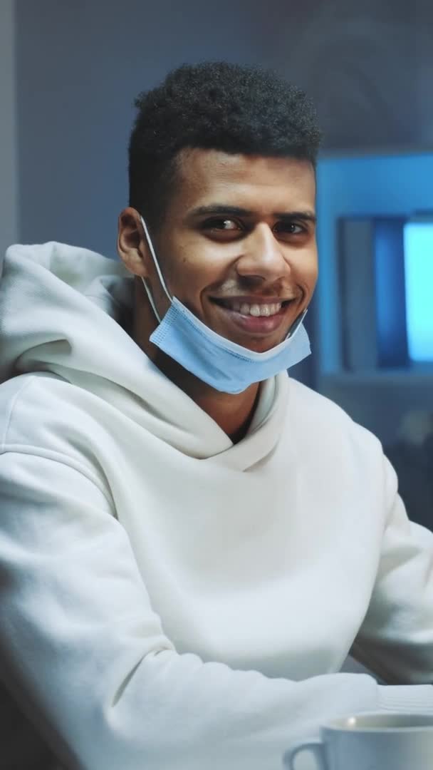 Hombre negro guapo con máscara médica sonriendo a la cámara mientras trabaja en casa — Vídeos de Stock