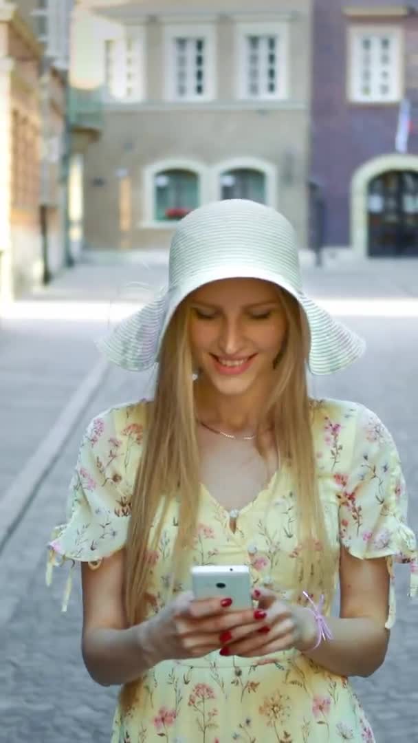 길거리에서 문자하는 여자. 하얀 모자를 쓴 쾌활 한 젊은 숙녀가구 시가지를 걷고 스마트폰으로 문자 메시지를 보내고 있다. — 비디오