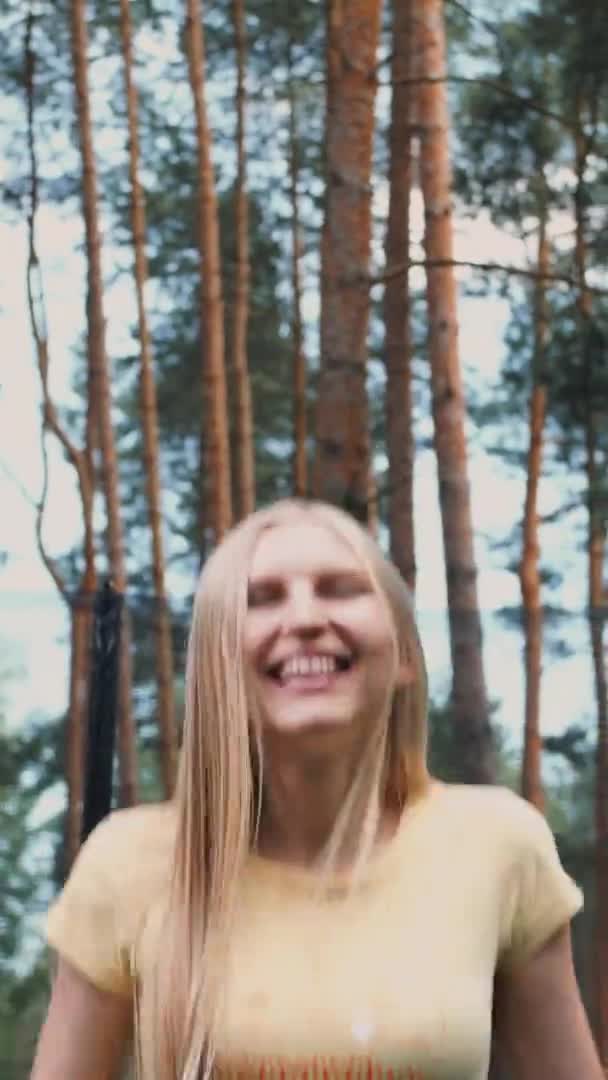 Salto feminino feliz no trampolim. Linda jovem loira descalça mulher em camisa de verão leve e jeans olhando para a câmera e sorrindo pulando em grande trampolim com árvores altas ao redor . — Vídeo de Stock
