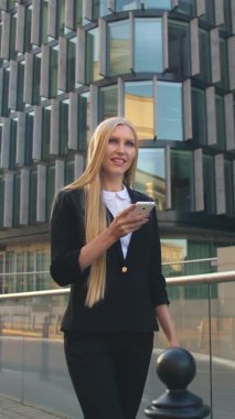 Modern woman walking on contemporary urban street. Lass using phone and walking on paved street of contemporary city with new buildings of offices in sunlight.
