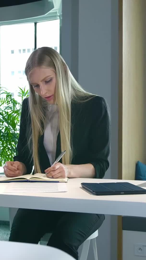 Mujer elegante que trabaja en una oficina elegante. Mujer rubia moderna en traje de moda sentada en la mesa en la oficina contemporánea de la luz y la escritura en papeles . — Vídeo de stock