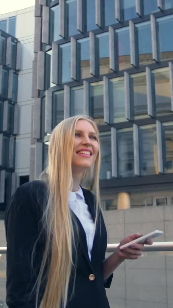 Mujer de negocios sonriente con teléfono en la calle. Chica rubia moderna en traje elegante sosteniendo el teléfono y de pie en la calle con edificios arquitectónicos modernos . — Vídeo de stock