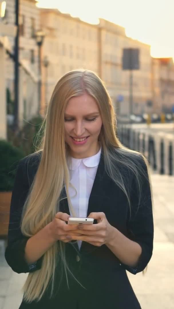 Mujer formal de negocios caminando por la calle. Elegante mujer rubia en traje y caminando por la calle y navegando por el teléfono inteligente con sonrisa contra el fondo urbano . — Vídeos de Stock