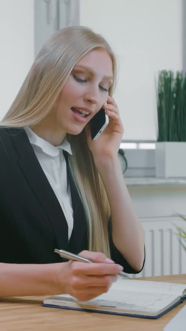 Geschäftsfrau im Büro im Smartphone-Gespräch. Elegante junge blonde Frau im Büroanzug sitzt am Arbeitsplatz und verhandelt per Handy in der Hand und schreibt die nötigen Informationen ins Notizbuch — Stockvideo