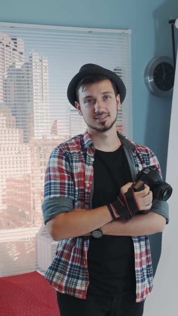 Guapo joven fotógrafo en sombrero sosteniendo la cámara y sonriendo en estudio moderno sesión de fotos — Vídeos de Stock