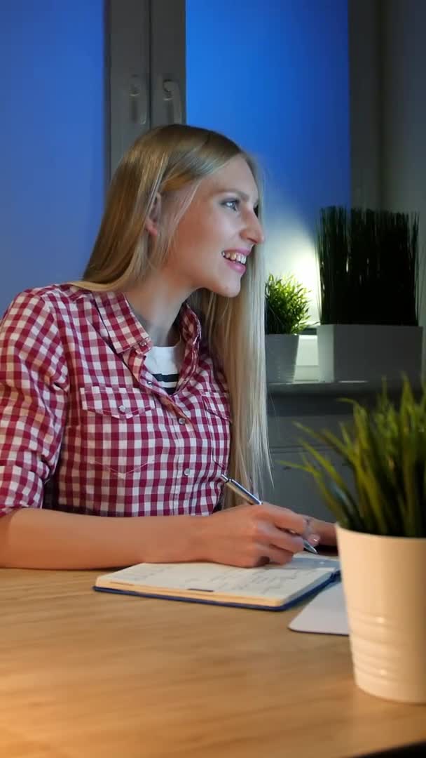 笑顔の女性は夜にコンピュータに取り組んでいます。小さなランプ木製の机に照らされて座っている小さなチェックされたシャツの女性を笑顔と興奮してコンピュータモニタを見て書き込み情報. — ストック動画