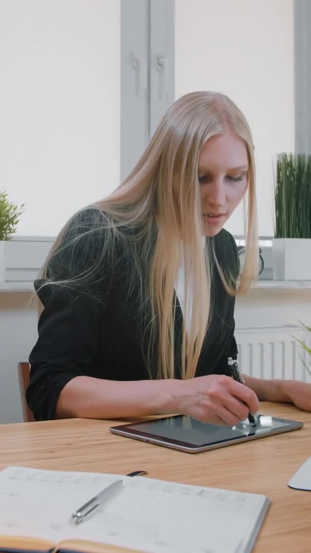 Femmes d'affaires travaillant sur tablette au bureau. Jolie femme blonde aux cheveux longs en costume élégant assis à un bureau en bois avec ordinateur et se concentrant sur le travail sur tablette avec stylo dans un bureau léger . — Video