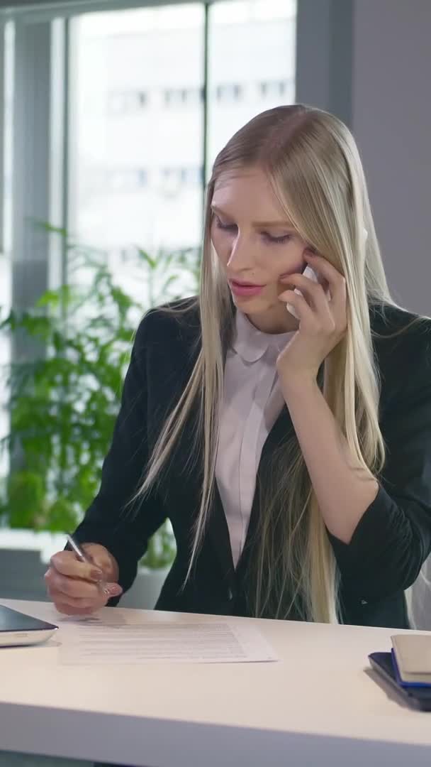 Модная деловая женщина смотрит ноутбук и разговаривает по телефону. Стильная молодая женщина, работающая в офисе за настольным ноутбуком и звонящая по телефону, улыбаясь . — стоковое видео