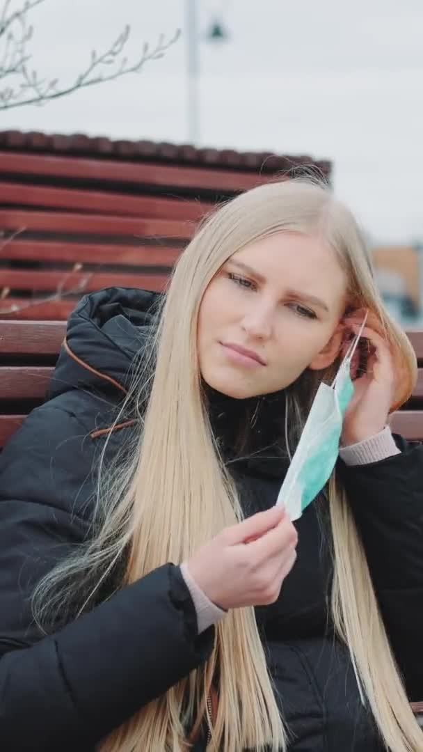 Молодая женщина сидит на скамейке и надевает защитную медицинскую маску на лицо — стоковое видео
