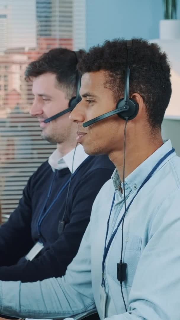 Portret van een lachende man van gemengd ras die een oproep aanneemt in een druk callcenter — Stockvideo