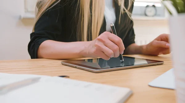 Donne d'affari che lavorano su tablet in ufficio. Attraente donna bionda dai capelli lunghi in abito elegante seduta alla scrivania in legno con computer e concentrata sul lavoro su tablet con penna in ufficio leggero . — Foto Stock