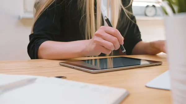 Geschäftsfrauen, die im Büro an Tablets arbeiten. Attraktive blonde Frau mit langen Haaren im eleganten Anzug sitzt am Holztisch mit Computer und konzentriert sich auf die Arbeit am Tablet mit Stift im hellen Büro. — Stockfoto