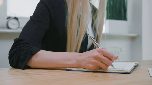 Γυναικεία γραφή με στυλό σε σημειωματάριο. Προβολή των καλλιεργειών της γυναίκας με μακριά ξανθά μαλλιά σε φωτεινό περιστασιακό πουκάμισο κάθεται στο ξύλινο γραφείο και γράφει πληροφορίες με γυαλιστερό μεταλλικό στυλό σε καθημερινό σχεδιασμό την ημέρα. — Φωτογραφία Αρχείου