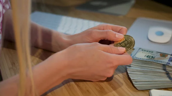 Female hands holding bitcoin. Crop view of hands of woman holding shiny metal bitcoin sitting at wooden desk with wad of cash nearby at night.