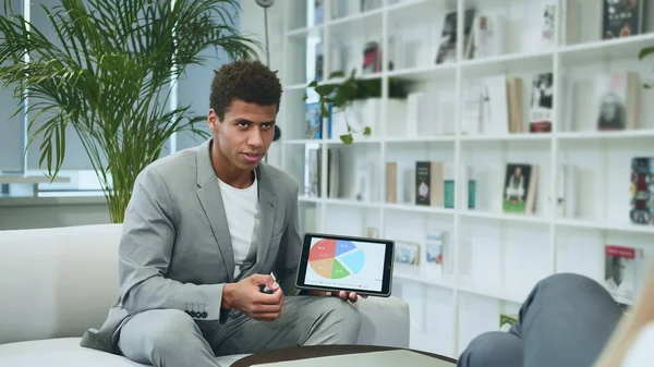 Black formal man making presentation with tablet. Young African-American man in suit sitting in office on sofa with anonymous colleague and making presentation with tablet.