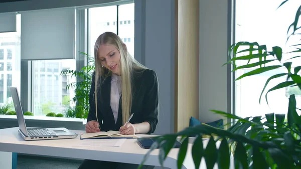 Mulher elegante trabalhando em escritório elegante. Mulher loira moderna em terno moderno sentado à mesa no escritório contemporâneo leve e escrevendo em papéis . — Fotografia de Stock