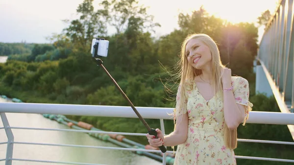 Fille prenant selfie sur le pont. Joyeux jolie femme blonde debout sur la passerelle piétonne et prendre selfie avec monopode au coucher du soleil . — Photo