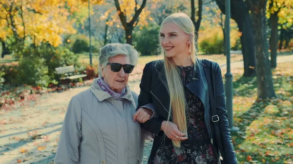 Old woman walking together with cheerful blonde girl in autumn park — Stock Photo, Image