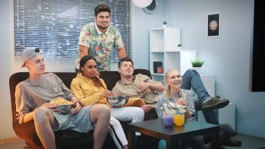 Heyecanlı arkadaşlar kanepede dinleniyor, TV izliyor ve evde atıştırmalık şeyler yiyorlar.