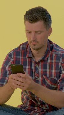 Kareli tişörtlü neşeli bir adam yerde oturmuş akıllı telefondan sohbet ediyor..