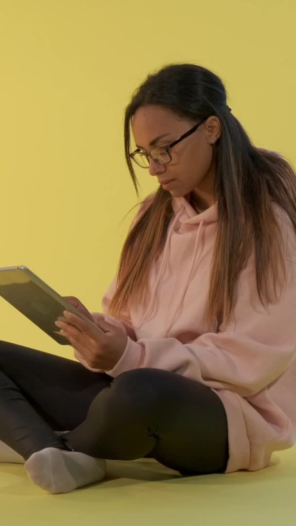 Atractiva chica de raza mixta leyendo algo en la tableta y luego mirando por encima de sus gafas a la cámara — Vídeos de Stock