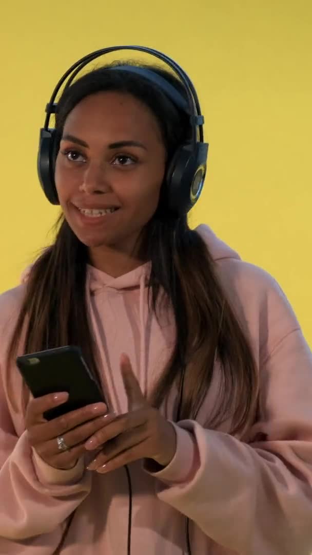 Glückliche multiethnische Frau, die mit Kopfhörern auf gelbem Hintergrund Musik hört — Stockvideo