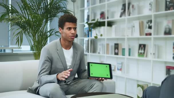 Černé formální člověk dělat prezentaci s tabletem s zelené monitory. Mladý americký muž v obleku, sedí v kanceláři na pohovce s anonymním kolegou a dělat prezentaci s tabletem. — Stock video
