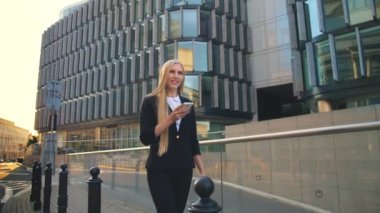 Modern woman walking on contemporary urban street. Lass using phone and walking on paved street of contemporary city with new buildings of offices in sunlight.