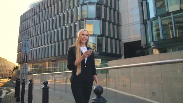 Moderne kvinde går på moderne bygade. Lass ved hjælp af telefon og gå på asfalterede gade i moderne by med nye bygninger af kontorer i sollys . – Stock-video