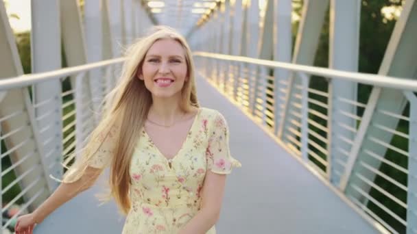 Uma mulher a olhar para trás na ponte. Menina loira bonita olhando para trás para a câmera enquanto corre na ponte com cabelo ondulado . — Vídeo de Stock