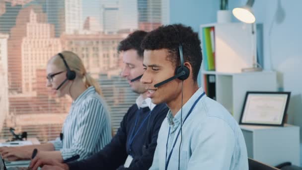 Close-up shot van een lachende man van gemengde rassen die een oproep aanneemt in een druk callcenter — Stockvideo