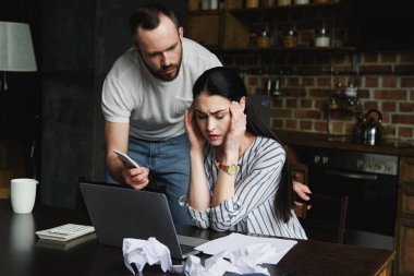 laptop ile masada oturan genç kadın vurguladı ve kızgın kocası onun smartphone ekran gösterilen süre kağıtları buruşuk