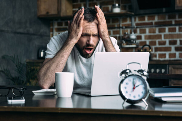 portrait of overworked freelancer remote working at table with laptop, clock and cup of coffee in kitchen at home