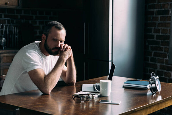 depressed young man sitting on kitchen with laptop and looking away