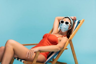 Güneş gözlüklü ve tıbbi maskeli bir kadın mavi güvertede tek başına oturuyor.