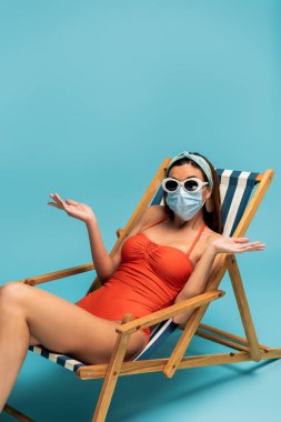 Güneş gözlüklü ve tıbbi maskeli kafası karışmış bir kadın mavi arka planda, iskemlede oturuyor.
