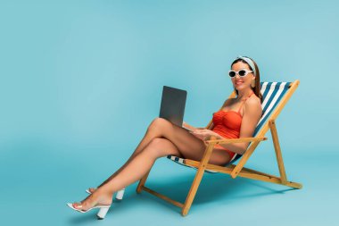 Freelancer with crossed legs and laptop smiling on deckchair on blue clipart