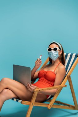 Freelancer in medical mask with laptop showing bottle of hand sanitizer on deckchair on blue clipart