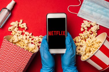 KYIV, UKRAINE - 26 Mart 2020: Patlamış mısır ve kırmızı zemin üzerinde el dezenfektanı bulunan netflix uygulamalı akıllı telefonu elinde bulunduran kişinin üst görüntüsü