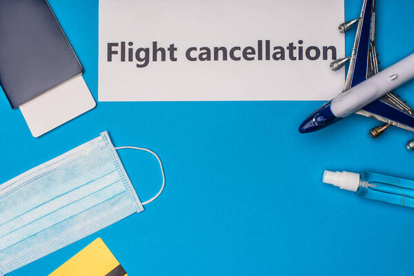 Top view of card with flight cancellation lettering near medical mask, toy airplane, passport and air ticket on blue background